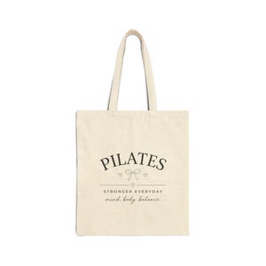 May include: A cream-colored canvas tote bag with two shoulder straps. The front features black text that reads PILATES, STRONGER EVERYDAY, and mind. body. balance. A small line drawing of a bow and three tiny hearts are centered below the word PILATES.