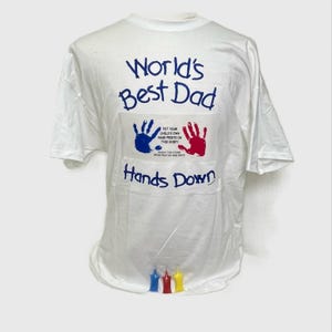 Puede incluir: Camiseta blanca con huellas de manos azules y rojas y el texto "World's Best Dad Hands Down". La camiseta también tiene el texto "Put your child's own hand prints on this shirt!" y tres pequeñas figuras de colores en la parte inferior.