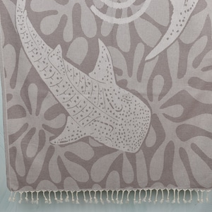 May include: A gray beach towel with a white whale shark design. The towel features a repeating pattern of abstract shapes and a fringe along the bottom edge. The whale shark is depicted in detail, with spots and markings.