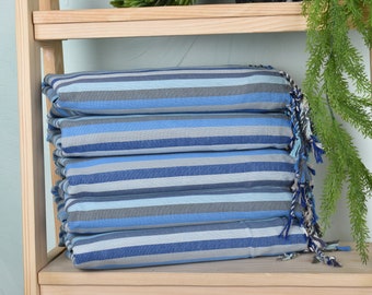 May include: A stack of five blue and gray striped towels with fringe on the ends. The towels are folded and stacked on a wooden shelf.