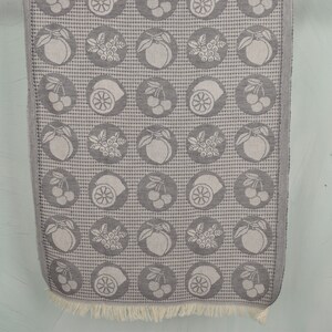 May include: A gray and white kitchen towel with a repeating pattern of lemons, cherries, and berries. The towel has a fringe border.