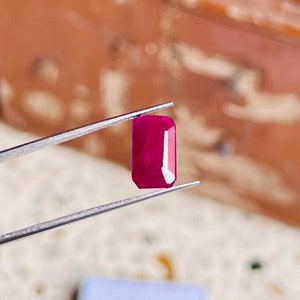 May include: A vibrant pink rectangular gemstone held by tweezers. The gemstone is smooth and polished, with a slight shimmer.