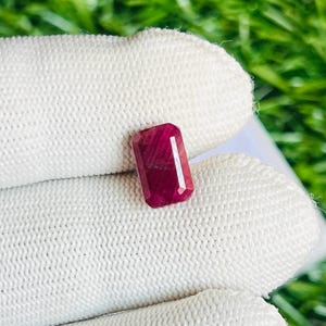 May include: A deep red gemstone with a rectangular shape, cut into a faceted emerald shape. The gemstone is displayed on a white fabric background.