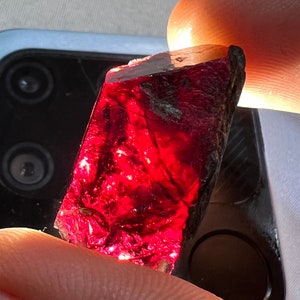 Op de afbeelding: Een diep rode, gefacetteerde edelsteen met een ruwe, natuurlijke textuur. De edelsteen wordt in een hand gehouden, en het licht reflecteert van zijn oppervlak.
