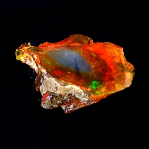 Puede incluir: Una gema de ópalo natural en bruto con un brillo iridiscente naranja, rojo y verde. El ópalo está engastado en una matriz de roca gris y marrón.