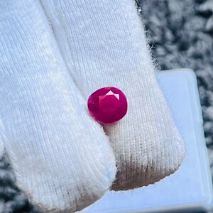 May include: A vibrant pink gemstone, likely a ruby, is displayed on a white surface. The gemstone is oval-shaped and has a smooth, polished finish.