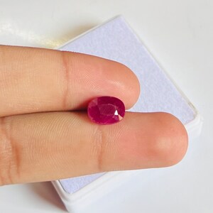 Puede incluir: Una piedra preciosa ovalada de color rojo oscuro, probablemente un rubí, se sostiene entre dos dedos.