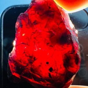 May include: A large, deep red, rough-cut gemstone with a slightly irregular shape. The gemstone is translucent and has a glassy luster.