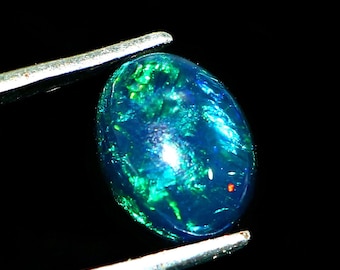 May include: An oval-shaped black opal gemstone featuring a cabochon cut with vibrant flashes of blue and green iridescence throughout the surface. The stone is held by silver-toned metal tweezers against a solid black background.