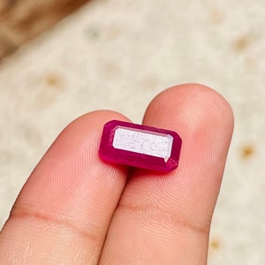 May include: A vibrant pink gemstone with a rectangular cut. The gemstone is held between two fingers.