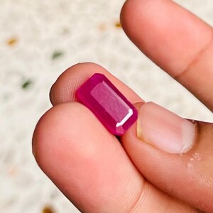May include: A vibrant, rectangular-cut gemstone with a deep magenta-pink hue. The stone features a faceted surface that reflects light, showcasing a clear, polished finish with sharp, defined edges and a consistent, saturated color throughout.