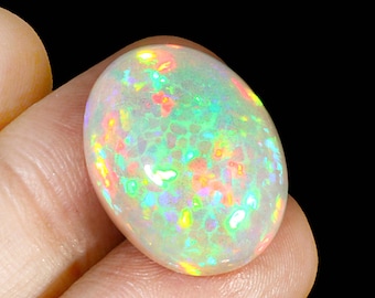 May include: An oval-shaped opal gemstone with a milky white base color. The opal displays a vibrant play-of-color, with flashes of green, yellow, orange, and red throughout its surface. The gemstone is held between fingers against a black background.