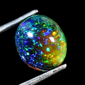 May include: A vibrant, oval-shaped opal gemstone held by silver tweezers. The opal displays a mesmerizing play of color, with flashes of blue, green, yellow, and orange against a dark background. The gemstone's surface is smooth and polished, reflecting light.