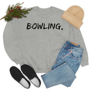 May include: Gray sweatshirt with the text "BOWLING." in black lettering. The sweatshirt is styled with blue jeans, a beige beanie, and black sneakers.