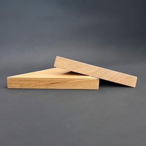 Può includere: Due blocchi espositivi in legno. Uno è un prisma triangolare, l'altro un prisma rettangolare. Entrambi sono in legno chiaro con venature visibili. I prismi sono disposti su uno sfondo grigio scuro.