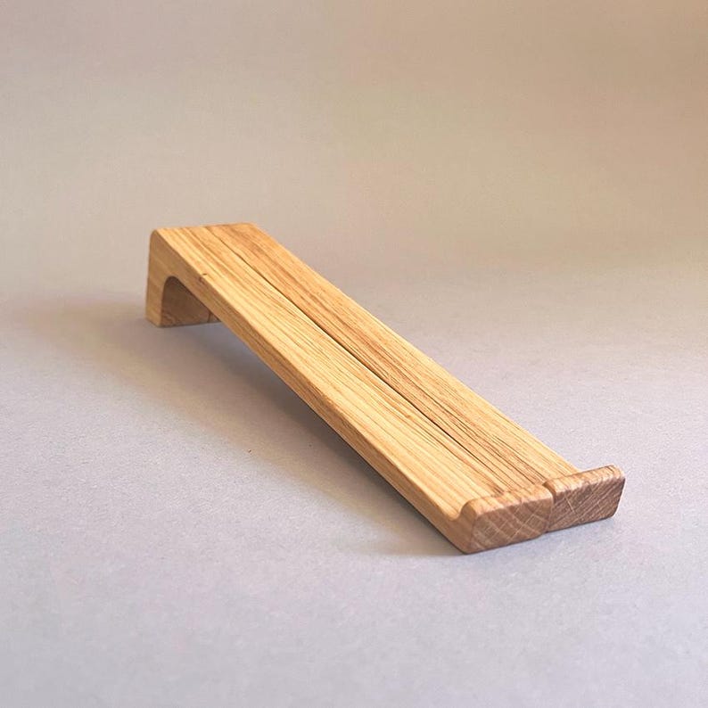 May include: A wooden tablet stand with a natural finish. The stand is angled, with a raised lip at the bottom and a curved support at the top. The stand is made of light-colored wood and has visible grain patterns.