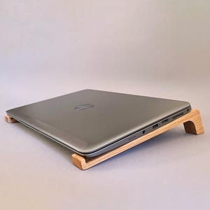May include: A gray HP laptop rests on a wooden laptop stand. The stand is made of light-colored wood and elevates the laptop at a slight angle. The laptop is closed, displaying the HP logo on the lid. The stand is designed to improve ergonomics.