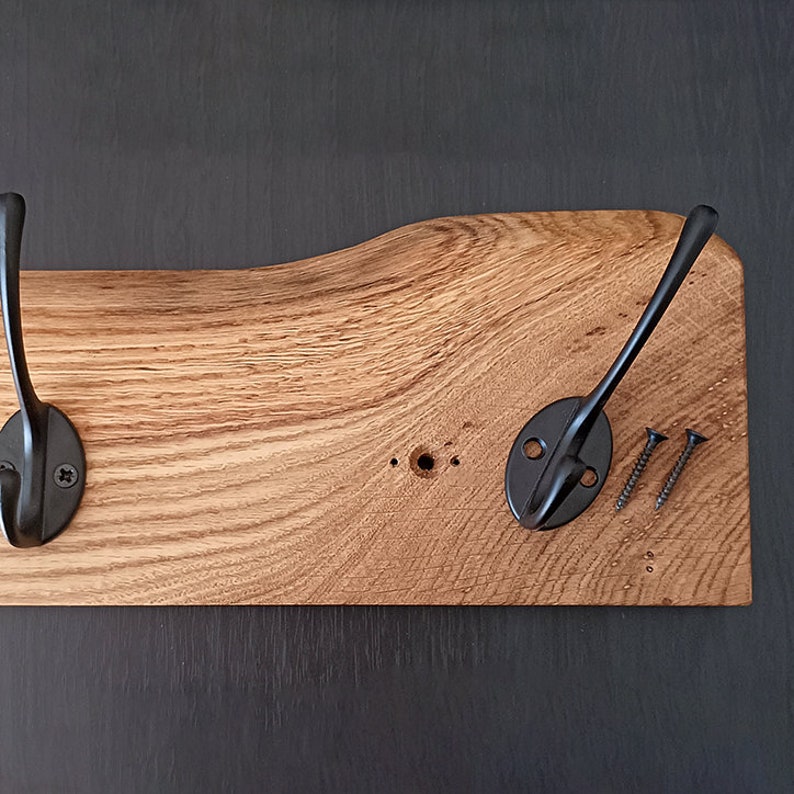 May include: A wooden coat rack with two black metal hooks. The wood has a natural grain and is unfinished. The hooks are attached with screws.