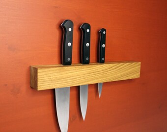 Peut inclure: Un porte-couteaux magnétique en bois avec trois couteaux attachés. Le porte-couteaux est en bois brun clair et est monté sur un mur rouge. Les couteaux ont des manches noirs et des lames argentées.