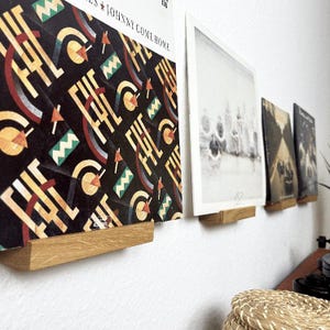 Könnte beinhalten: Vier Schallplatten werden an einer weißen Wand mit Hilfe von Holzregalen präsentiert. Die Schallplatte links hat ein schwarz-weißes geometrisches Muster mit dem Text "JOHNNY COME HOME". Die anderen Schallplatten sind Schwarzweißfotos.