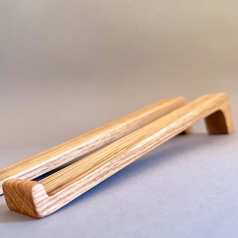May include: Two wooden cabinet pulls with a natural wood grain finish. The pulls have a rectangular shape with a curved handle, and are designed for easy gripping. The pulls are made of light-colored wood.