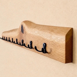 Peut inclure: Un porte-manteau mural composé d'une planche en bois clair naturel présentant un nœud et un grain apparents. Le support est équipé d'une série de crochets en métal noir courbés, fixés régulièrement sur le bord avant pour suspendre des objets.