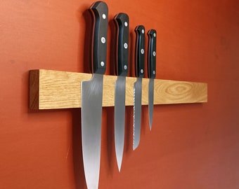 Peut inclure: Quatre couteaux de cuisine à manche noir avec des lames argentées sont montés sur une bande magnétique en bois. Les couteaux sont disposés par ordre de taille, le plus grand couteau à gauche et le plus petit couteau à droite.