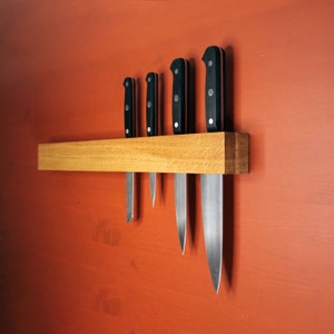 Op de afbeelding: Vijf keukmessen met zwarte handvatten zijn gemonteerd op een magnetische houten strip. De messen zijn in een rij gerangschikt, met het grootste mes rechts en het kleinste mes links.