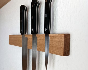 Peut inclure: Trois couteaux de cuisine avec des manches noirs sont montés sur un support magnétique en bois. Les lames sont argentées et le support en bois est de couleur marron clair. Les couteaux sont de tailles variables.