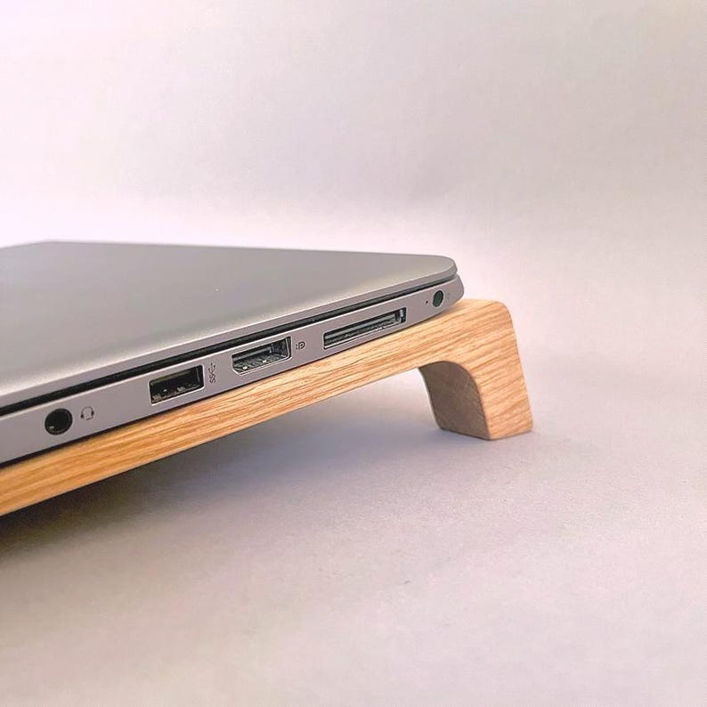 May include: A silver laptop rests on a light-colored wooden laptop stand. The stand has a curved design with a natural wood grain finish. The laptop's ports and side profile are visible, showcasing its sleek, modern design.
