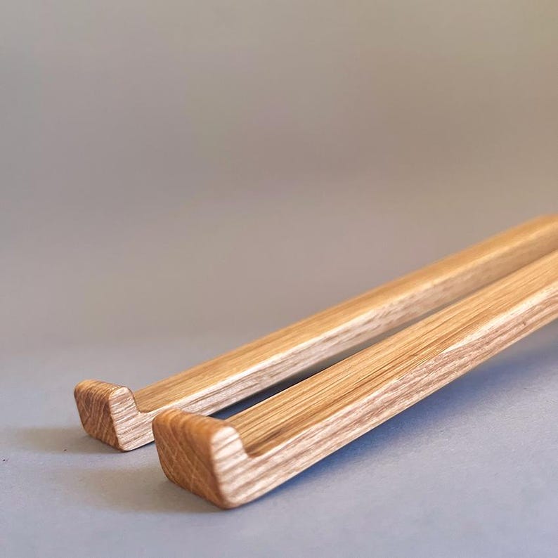 May include: Two wooden laptop stands with a light brown finish. The stands have a rectangular shape with a curved notch at one end. The wood grain is visible, and the stands are set against a gray background.