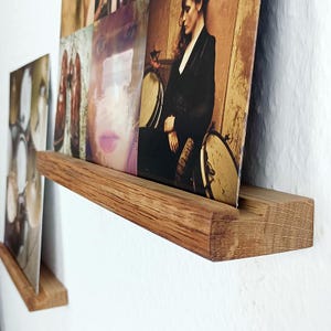 Może przedstawiać: Dwie drewniane półki ścienne z naturalnym wykończeniem drewna, zaprojektowane do przechowywania płyt winylowych. Półki są zamontowane na białej ścianie, a na każdej z nich leży płyta.