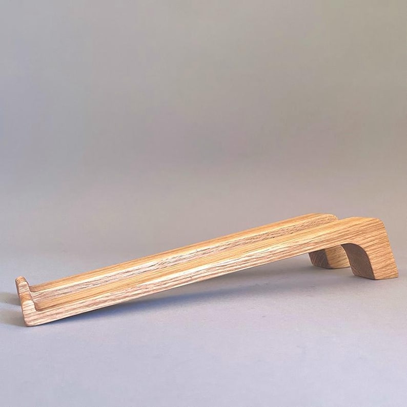 May include: A light-colored wooden laptop stand with a sloped design. The stand has a rectangular base and a curved support at the back. The wood grain is visible, and the stand is designed to elevate a laptop for ergonomic use.
