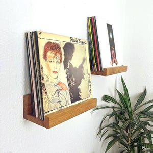 Może przedstawiać: Dwie drewniane półki zamontowane na białej ścianie, każda z płytami winylowymi. Górna półka ma jedną płytę z białą okładką i różowo-niebieskim wzorem. Dolna półka ma stos płyt z okładką przedstawiającą portret Davida Bowie. Tekst "David Bowie" i "Scary" jest widoczny na okładce.