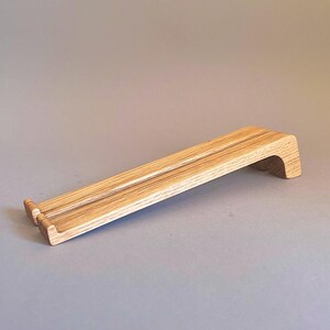 May include: A light-colored wooden laptop stand with a sloped design. The stand features a raised edge at the front and a curved support at the back. The wood grain is visible, and the stand is designed to elevate a laptop for ergonomic use.