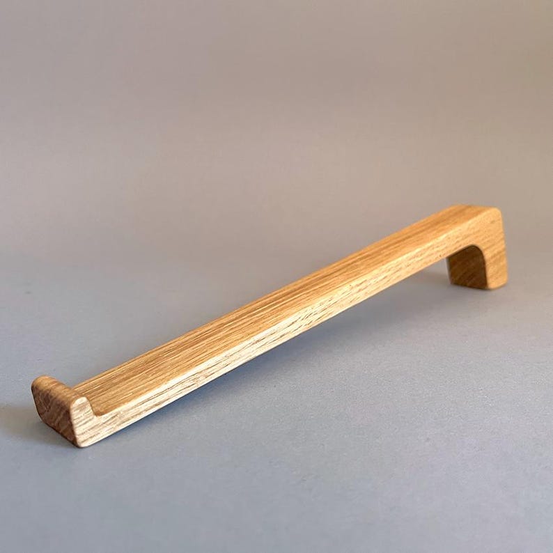 May include: A light-colored wooden cabinet pull with a rectangular bar and a curved end. The pull is made of natural wood and has a smooth finish. The cabinet pull is approximately 12 inches long.
