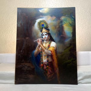 May include: A painting of Krishna, a Hindu deity, playing a flute. He is wearing a blue and yellow robe, a crown of feathers, and a necklace of beads. The background is a lush green forest with a waterfall.