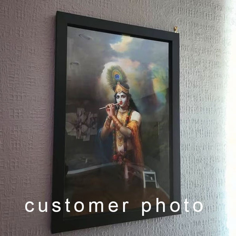 May include: A framed painting of a Hindu deity playing a flute. The deity is depicted with blue skin, a crown of feathers, and a yellow and orange robe. The painting is set against a cloudy sky background.