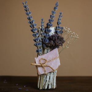 May include: A bundle of dried lavender, purple flowers, and white sage, tied with twine and a pink crystal. The lavender has blue-purple flower spikes. The sage is wrapped in a bundle. The background is a solid brown.