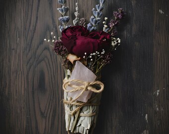Può includere: Un mazzo di salvia essiccata, rose rosse, lavanda e altri fiori secchi, legato con spago. Un cristallo rosa è inserito nel mazzo. Su uno sfondo di venature di legno scuro.