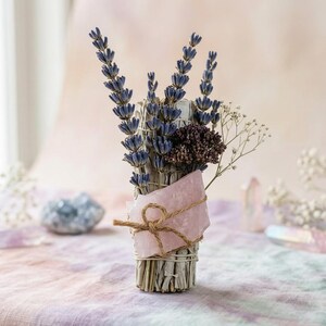 May include: A bundle of dried sage, lavender, and other dried flowers, tied with twine and a pink crystal. The arrangement is set on a pastel-colored surface, with additional crystals in the background. The sage bundle is approximately 6 inches tall.