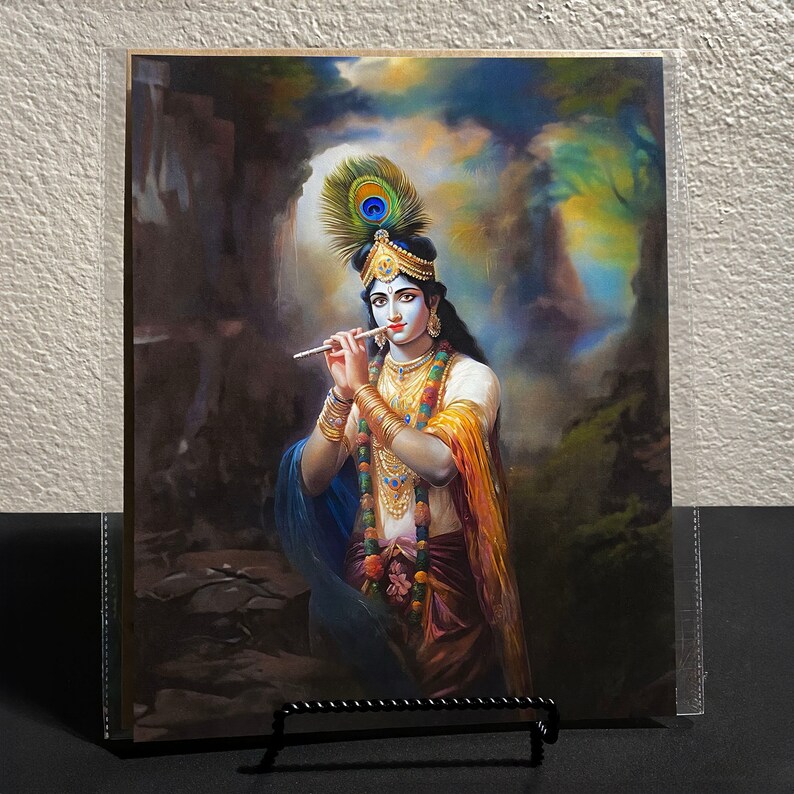 May include: A painting of Krishna playing a flute, with a peacock feather headdress, standing in a forest setting. He is wearing a yellow and blue robe, with gold jewelry and a garland of flowers.