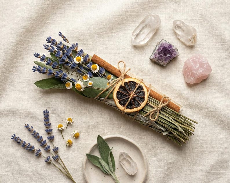May include: A collection of dried herbs, lavender, and chamomile flowers tied with twine, a dried orange slice, and cinnamon sticks. Clear quartz, amethyst, and rose quartz crystals are arranged on a neutral linen surface.