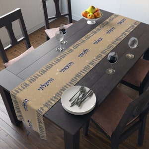 May include: A beige table runner with a repeating pattern of blue silhouettes of people and the Hebrew words "Shalom" and "Mazel Tov" printed on it. The runner is laid out on a dark brown wooden table with two wine glasses, a plate of fruit, and silverware.