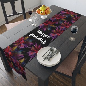 May include: A black table runner with a colorful powder explosion design and the text "Happy Diwali" in white. The runner is on a dark wood table with a bowl of apples, two wine glasses, and a plate with silverware.