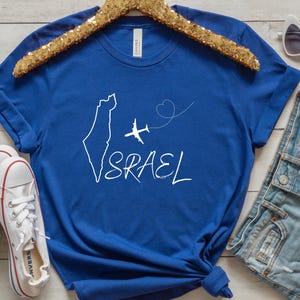 May include: A royal blue t-shirt with a white outline of Israel, a white airplane, and a heart with a dotted line. The text "ISRAEL" is written in white cursive.