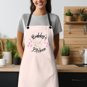 May include: A pink apron with black straps and the text "Bubby's Kitchen" in black with a pink bowl and whisk illustration.  Pink hearts are scattered around the text.
