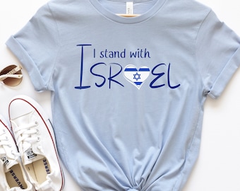 May include: Light blue t-shirt with the text "I stand with ISRAEL" in blue lettering. A heart with the Israeli flag is in the middle of the word ISRAEL.