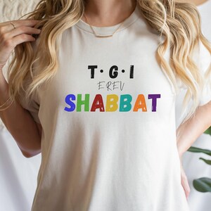 May include: White t-shirt with the text "T.G.I. EREV SHABBAT" in rainbow colors.