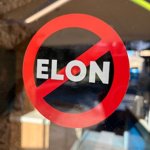 May include: A red circle with a diagonal line through it, with the word "ELON" in white letters inside. The circle is on a glass surface.